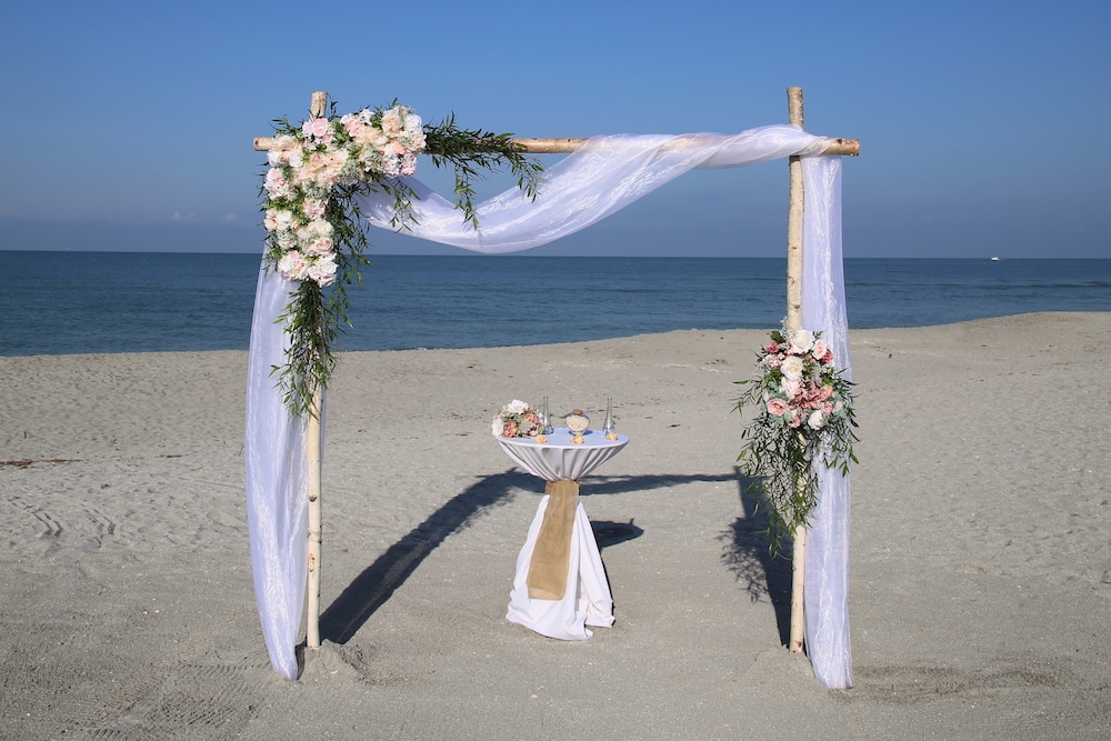 Beach Wedding Ceremony setup on Florida beach with blue skies. The arch is draped with white organza and asymmetrical flower arrangements.