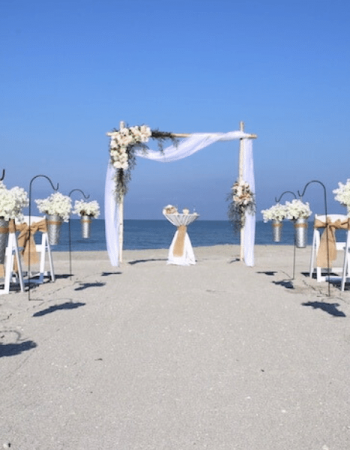 Beach Wedding Ceremony setup on Florida beach with blue skies. The arch is draped with white organza and asymmetrical flower arrangements.
